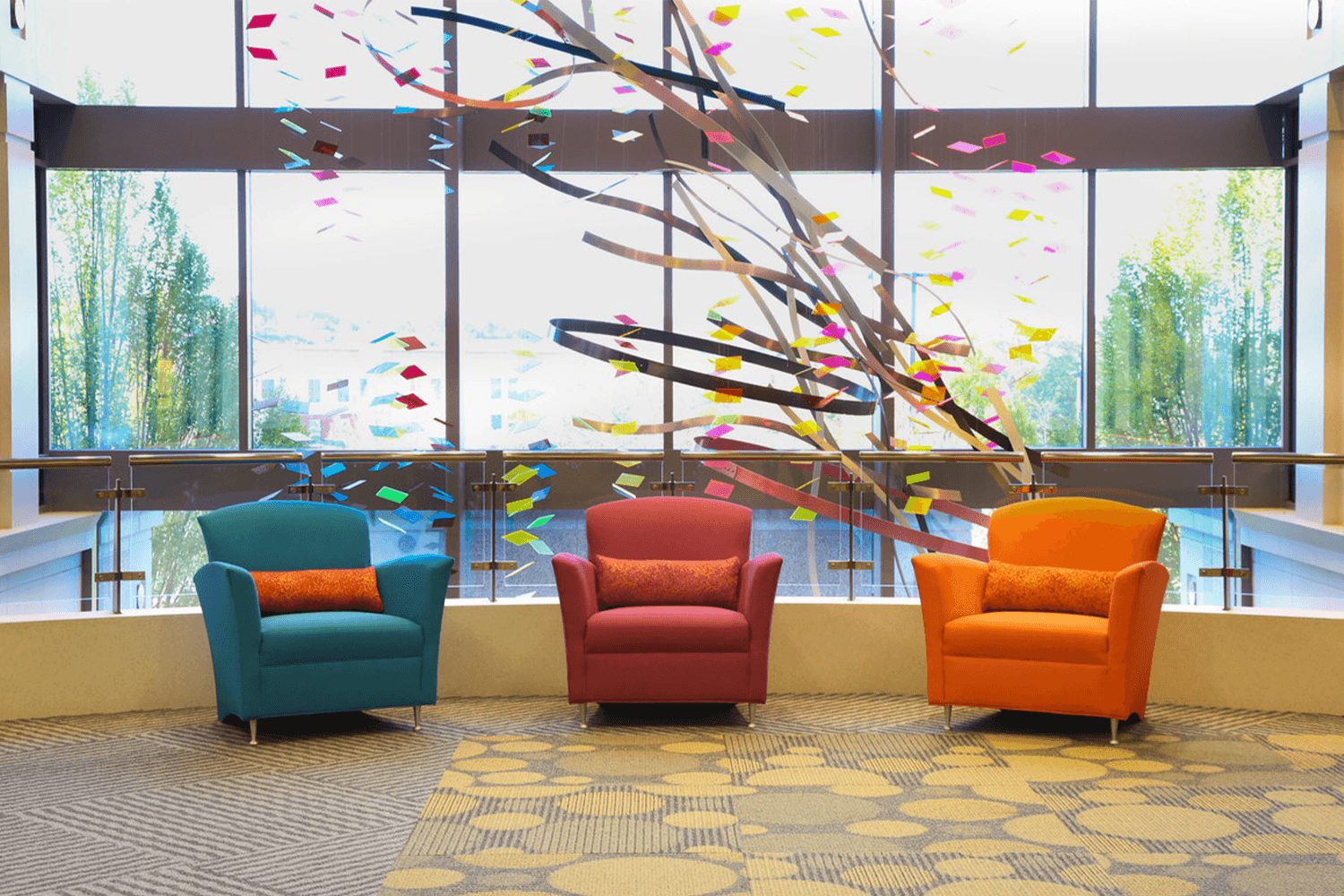 three vibrantly colored chairs, blue, light-red, and orange, overlooking a hanging sculpture and a wall fo windows