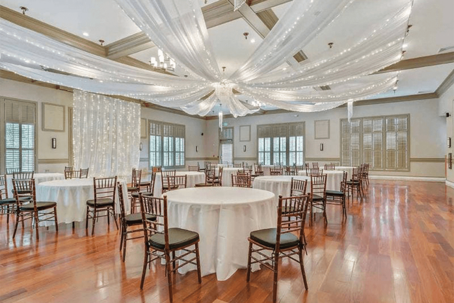 Event room with shiny wood floors, white drapery with string-lights, and tables with white table cloths