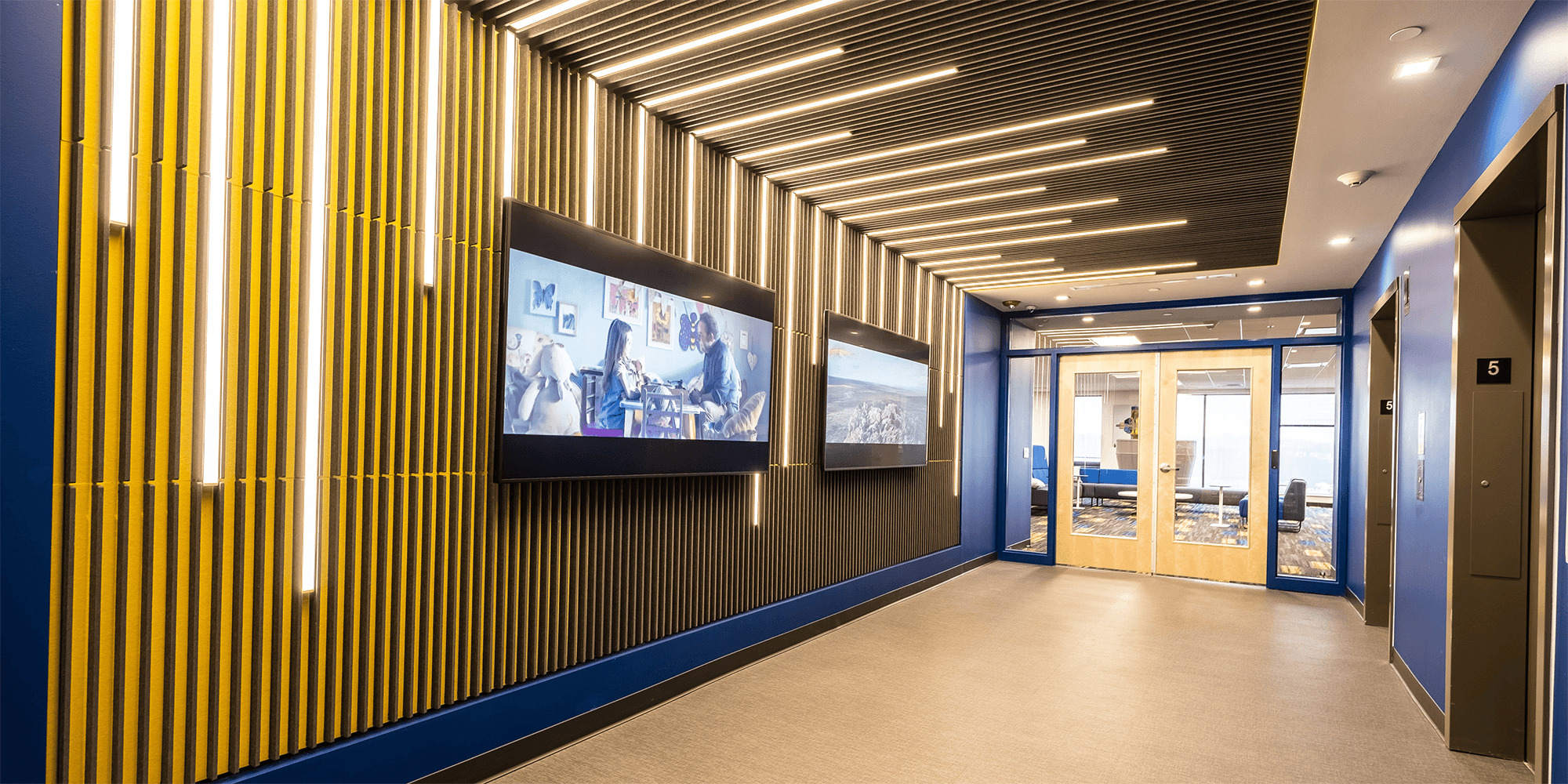 hallway with abstract blue and yellow walls, linear light fixtures, and two TVs