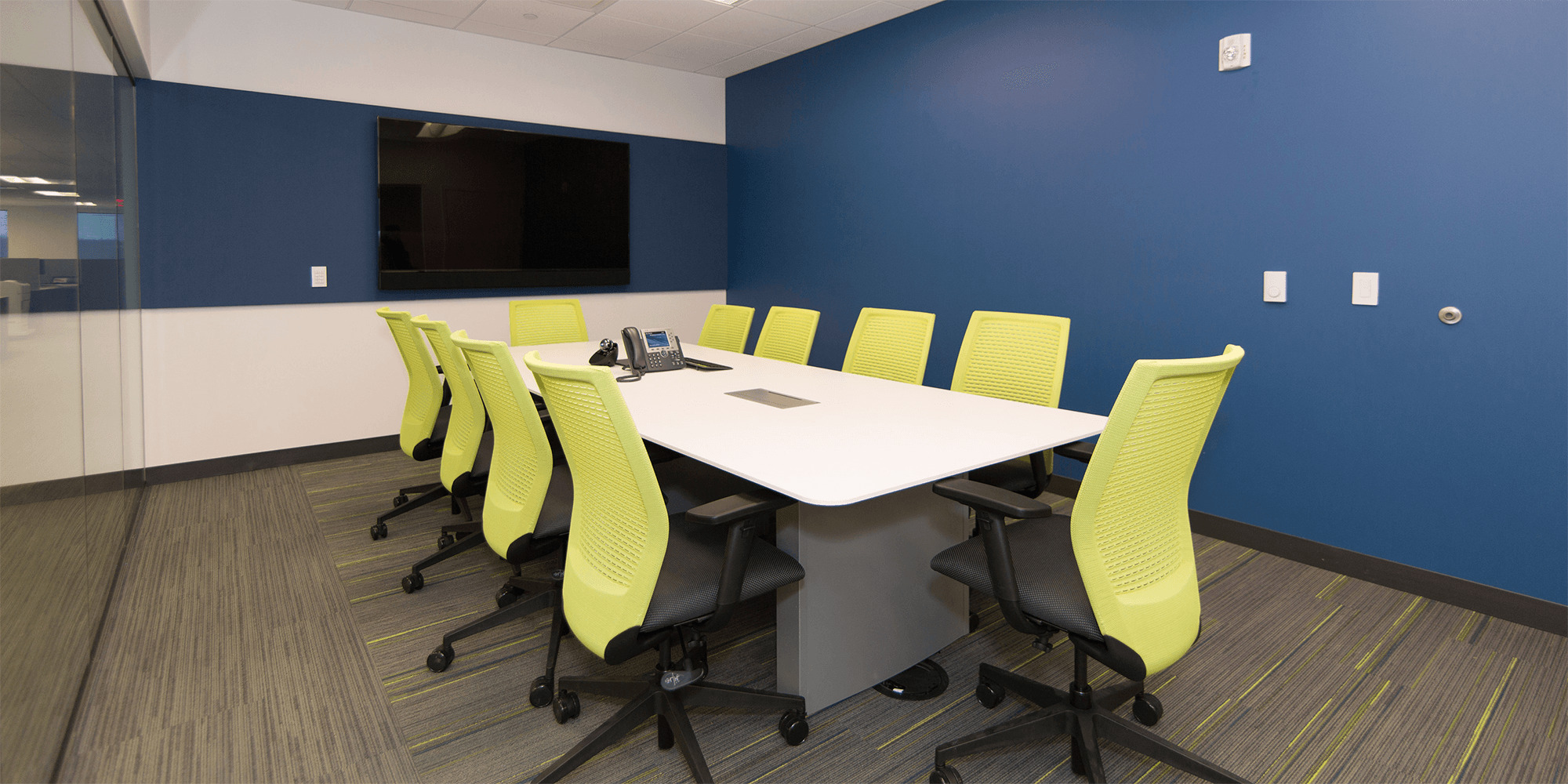a blue conference room with a glass wall, a TV, white tables, and neon green chairs